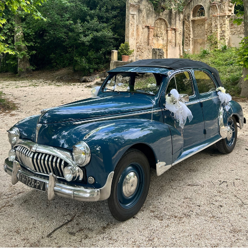 Voiture bleue rétro décorée de rubans blancs stationnée devant un lieu de cérémonie